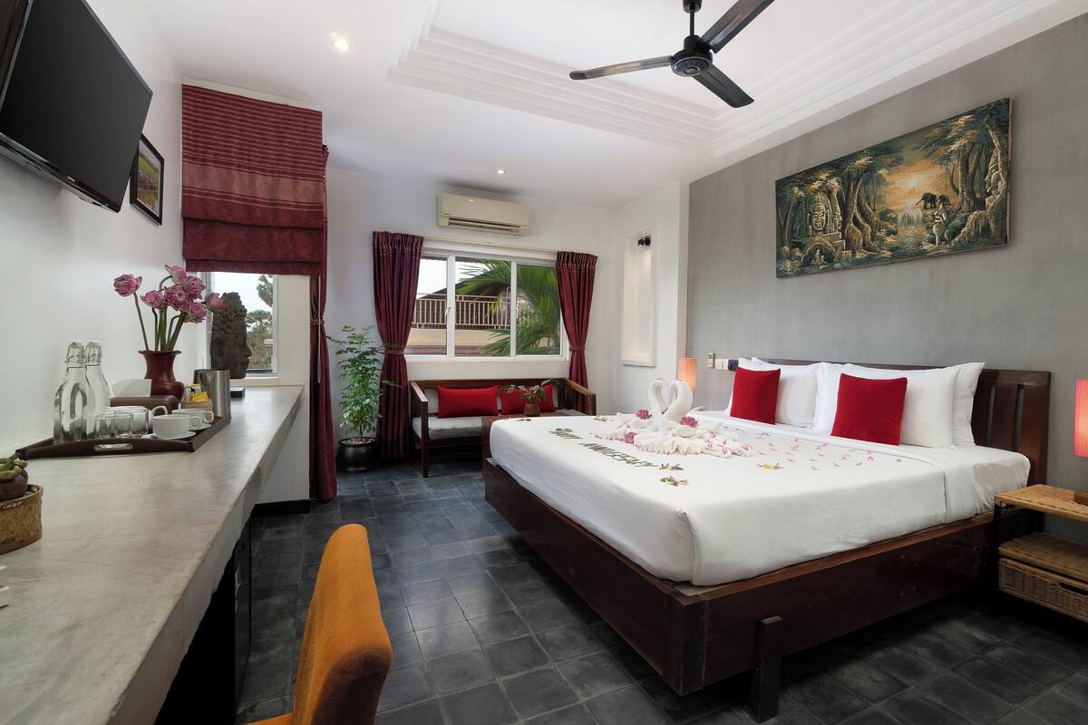 Habitación de hotel con cama, lámparas de noche, escritorio con televisor, ventana con cortinas y un cuadro en la pared.