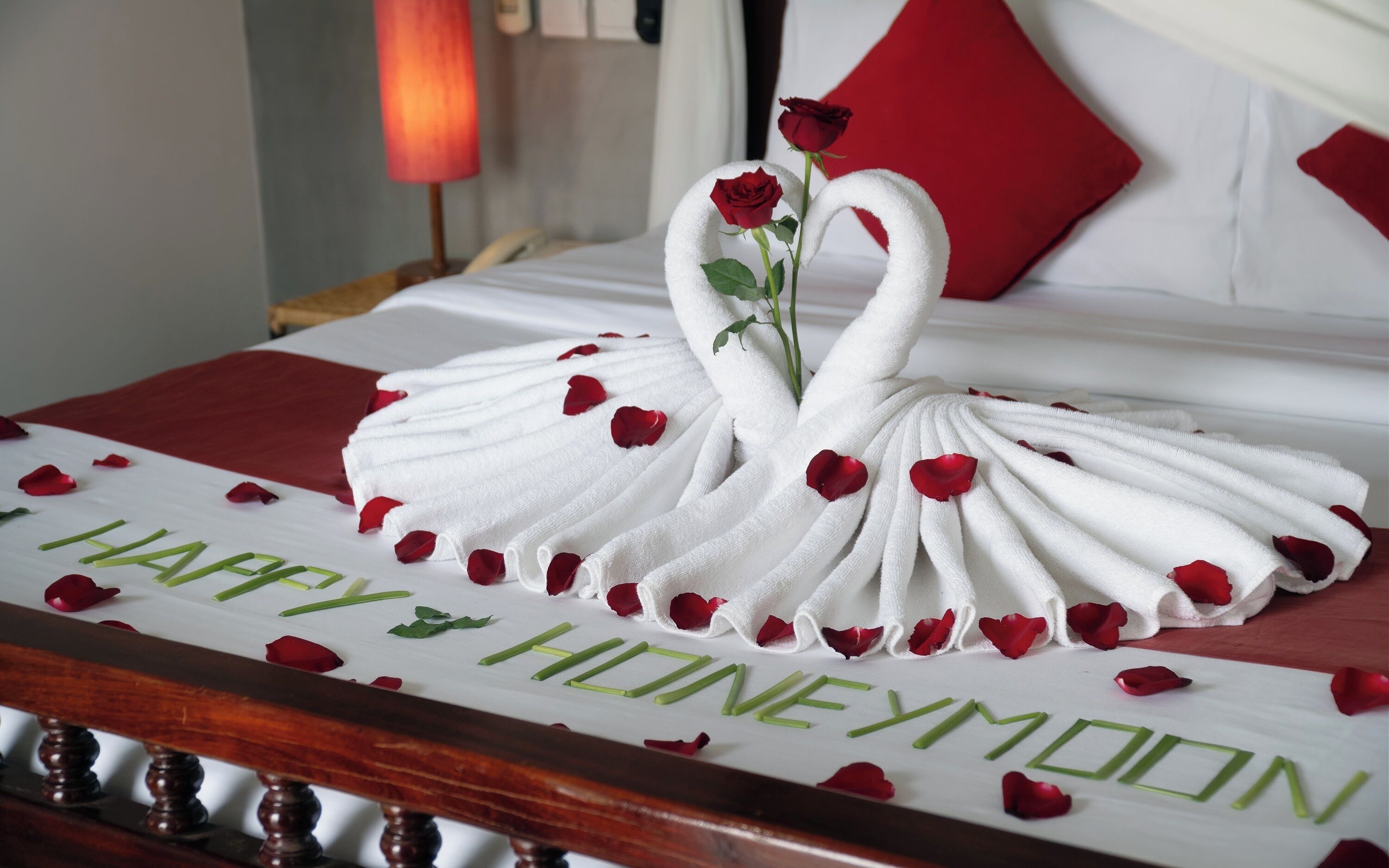 A bed with a towel arranged in the shape of swans, rose petals, and the words 'HAPPY HONEYMOON' on the bedspread.