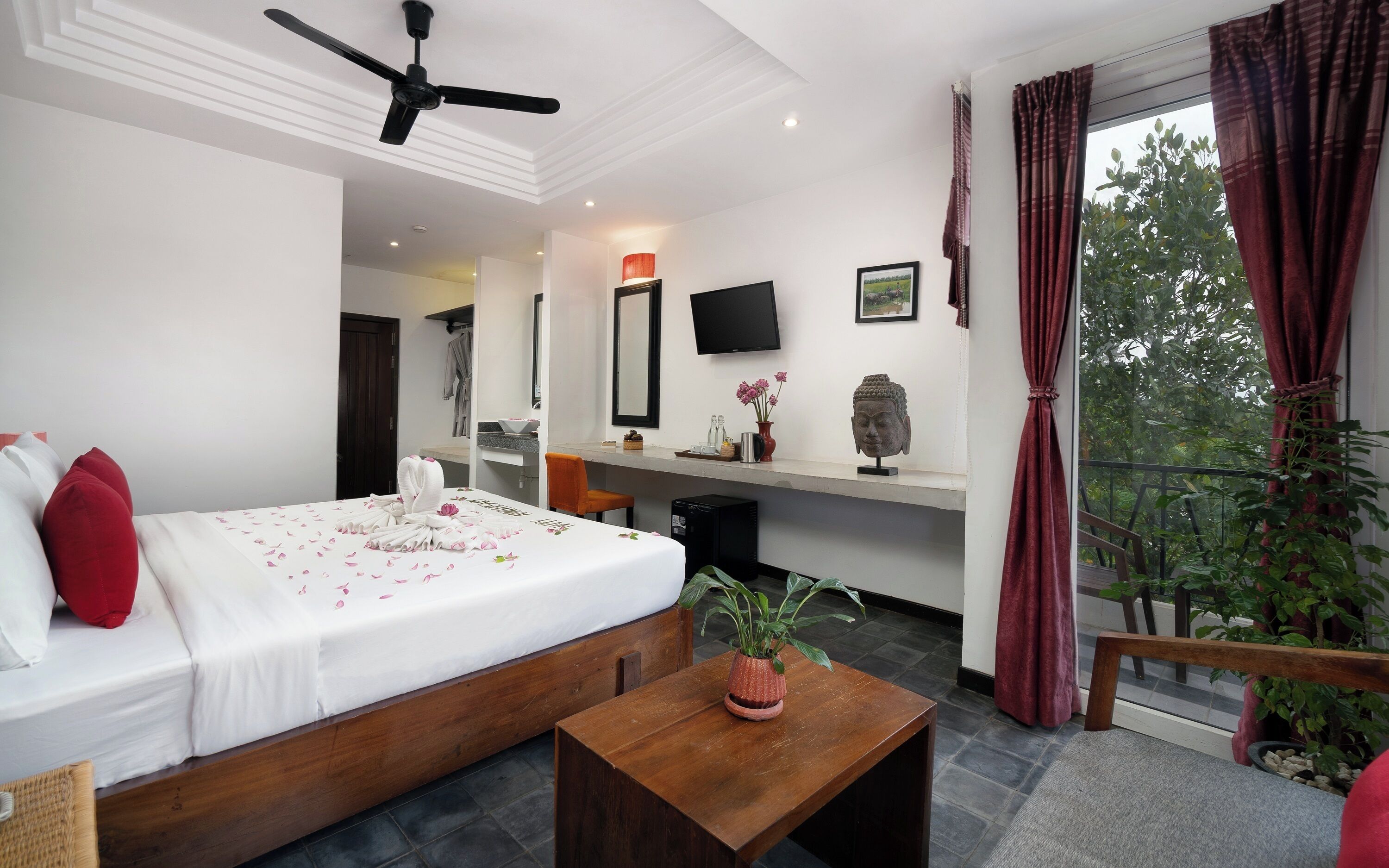 Ein Hotelzimmer mit Bett, Schreibtisch, Fernseher, einer Buddha-Statue und einem Balkon mit Pflanzen.