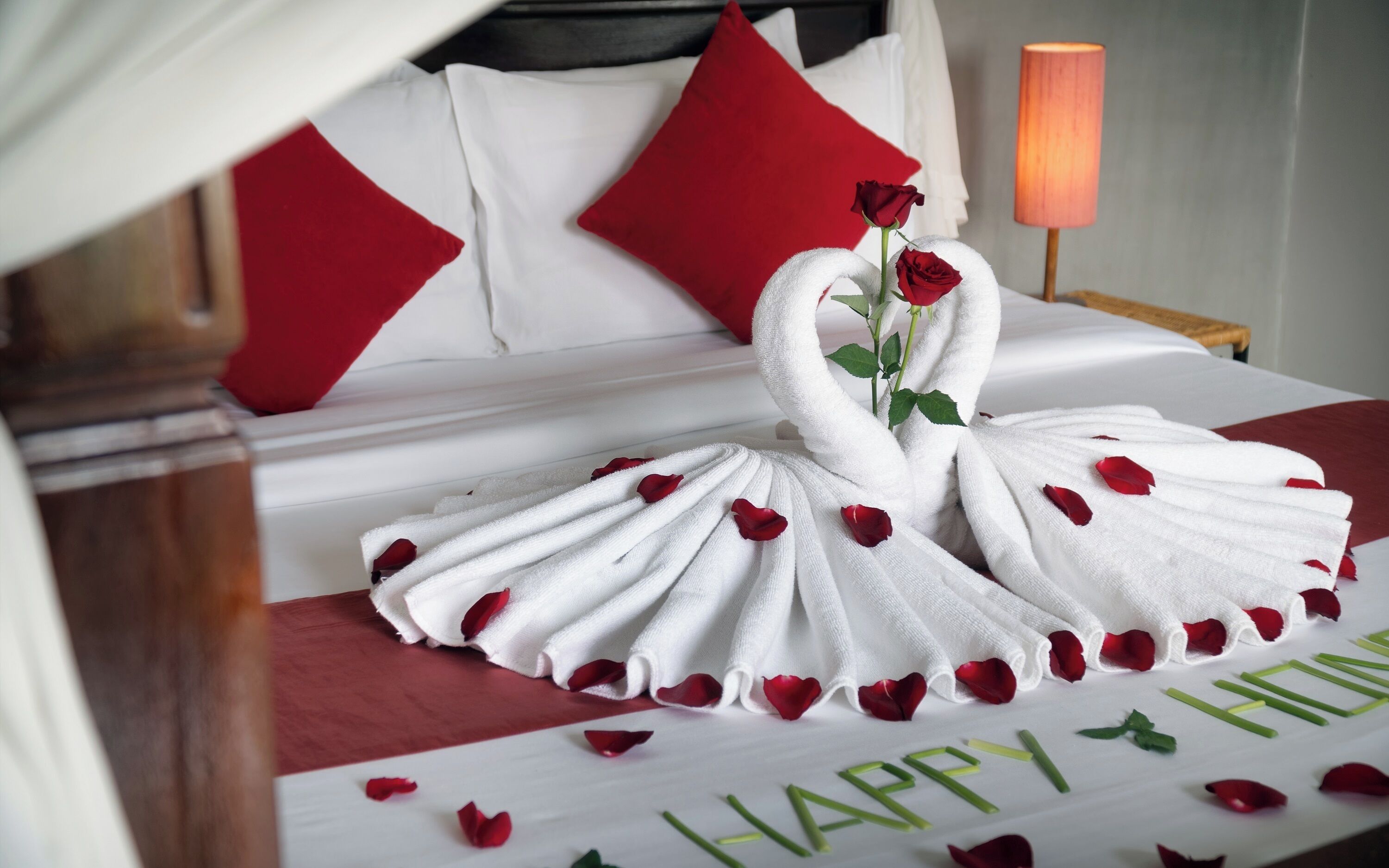 Una cama con una colcha blanca, almohadas rojas y una presentación de toallas en forma de rosa, con el mensaje 'Happy New Year'.