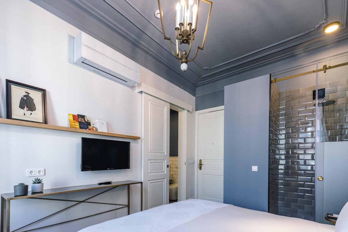 A modern bedroom with a large bed, a mounted TV, a small console table with decorative items, and a unique brick-patterned shower enclosure.