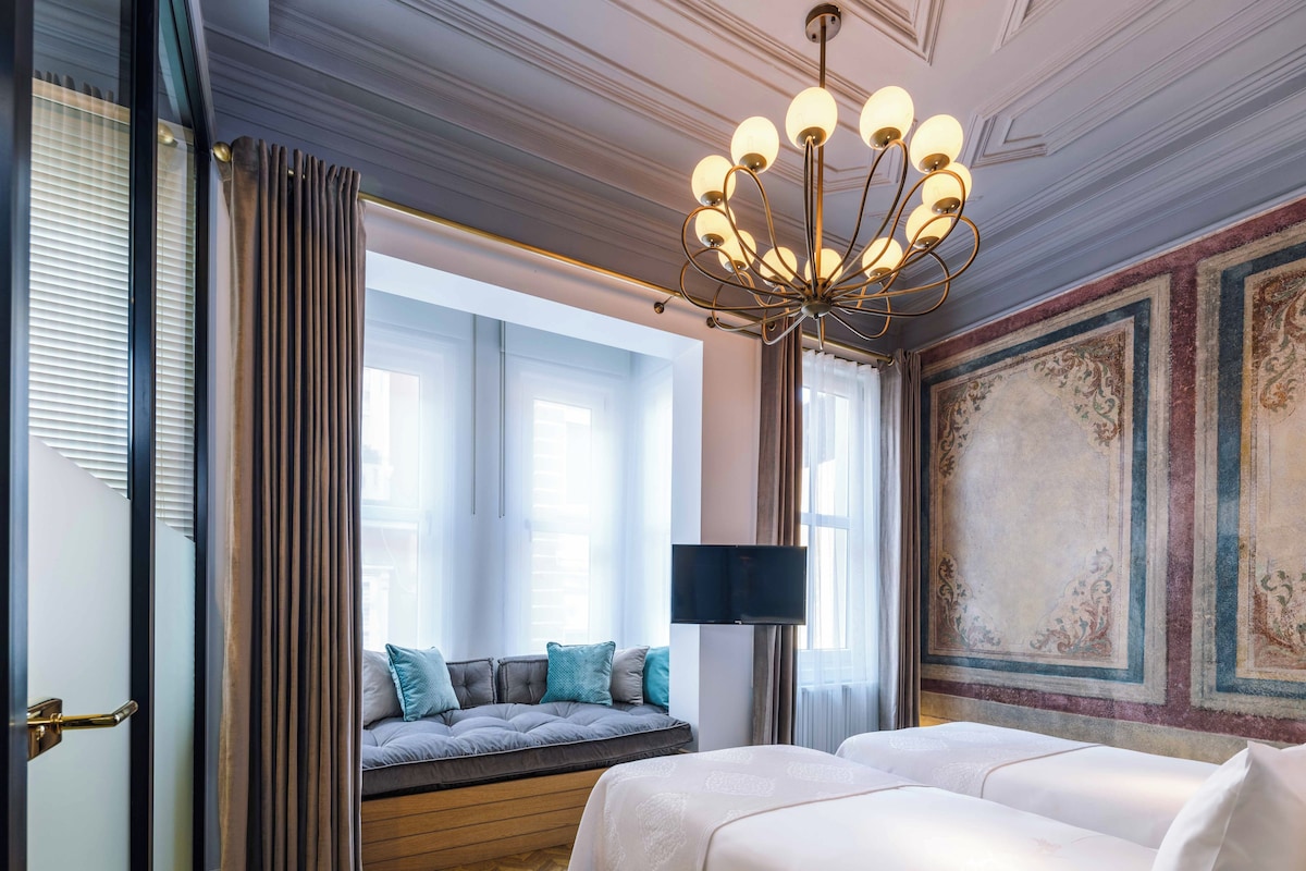 A bedroom with a large bed, a window seat, a chandelier, and a decorative wall.