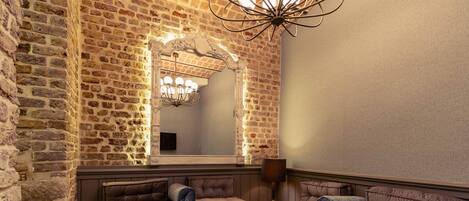 Um quarto aconchegante com parede de tijolos, um espelho grande e um lustre moderno. Há dois sofás e uma pequena mesa com objetos decorativos.