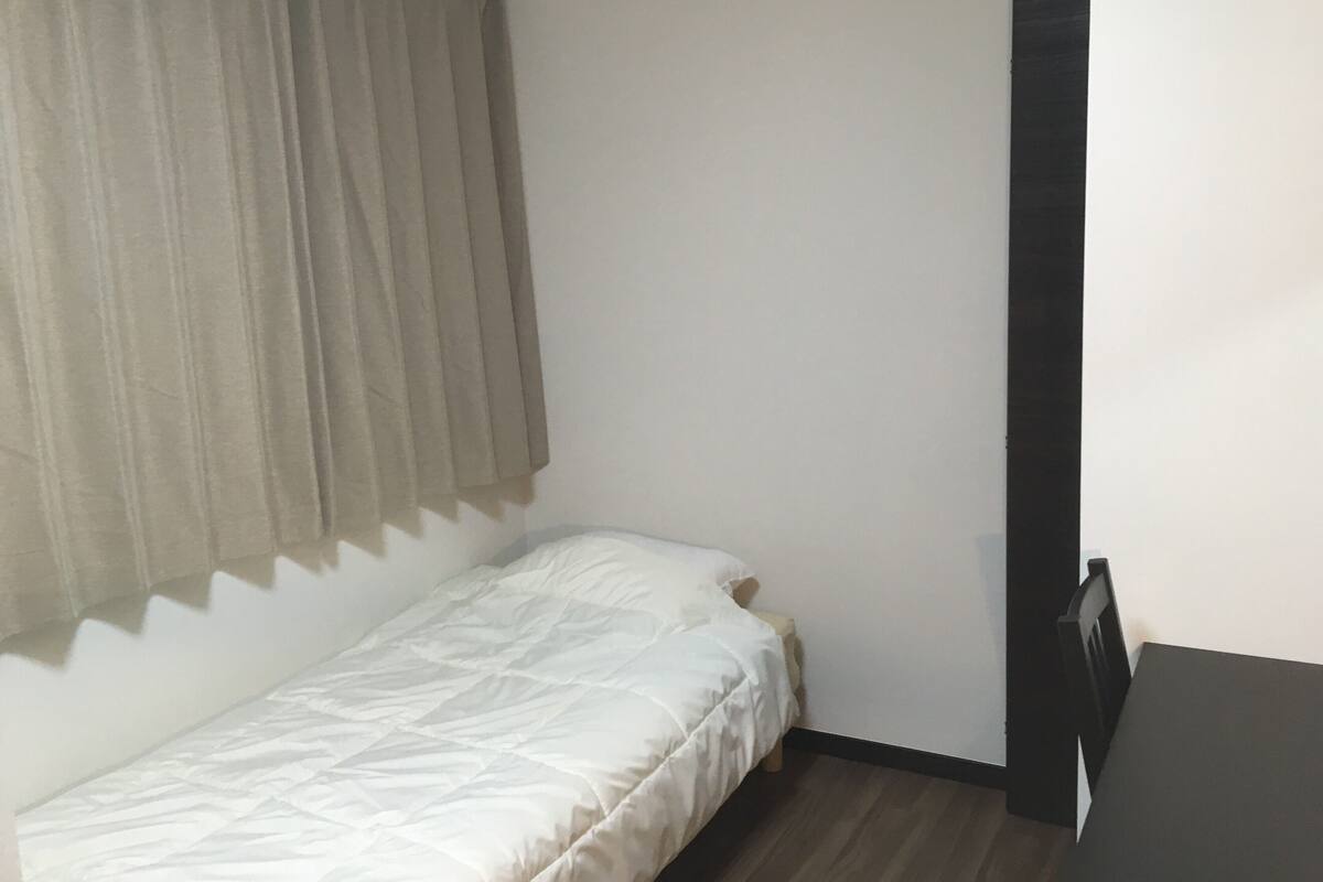 Ein kleines Zimmer mit einem Einzelbett, Holzboden, einem dunkelbraunen Tisch und einem Fenster mit cremefarbenen Vorhängen.