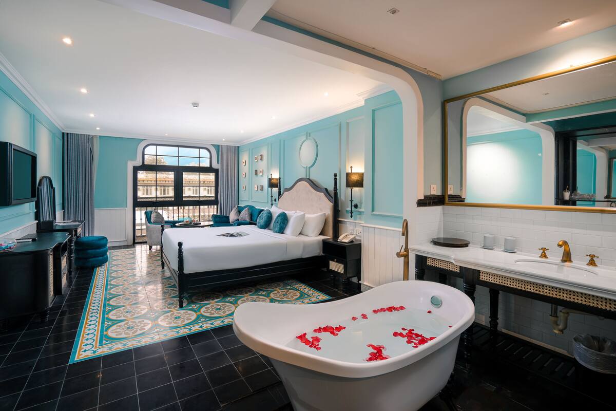 A modern hotel room with a large bed, a sitting area, a bathtub with rose petals, a bathroom with a large mirror, and a view of the city through the window.
