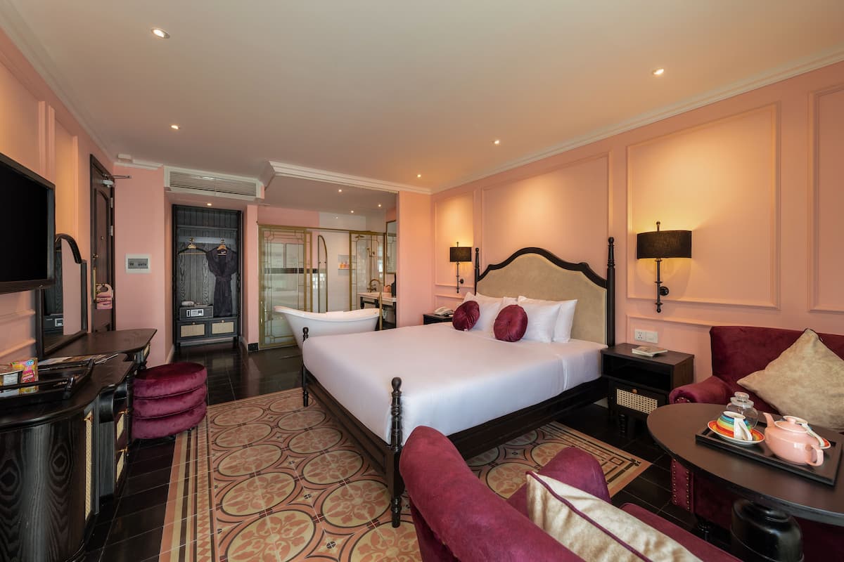 A hotel room with a large bed, a sitting area with a couch and armchair, a vanity with a mirror, a bathtub, and a television.