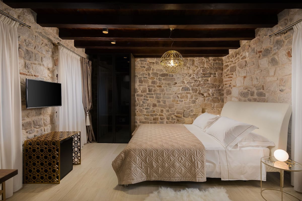 A bedroom with a stone wall, a bed with a quilted cover, a television mounted on the wall, and a hanging light fixture.
