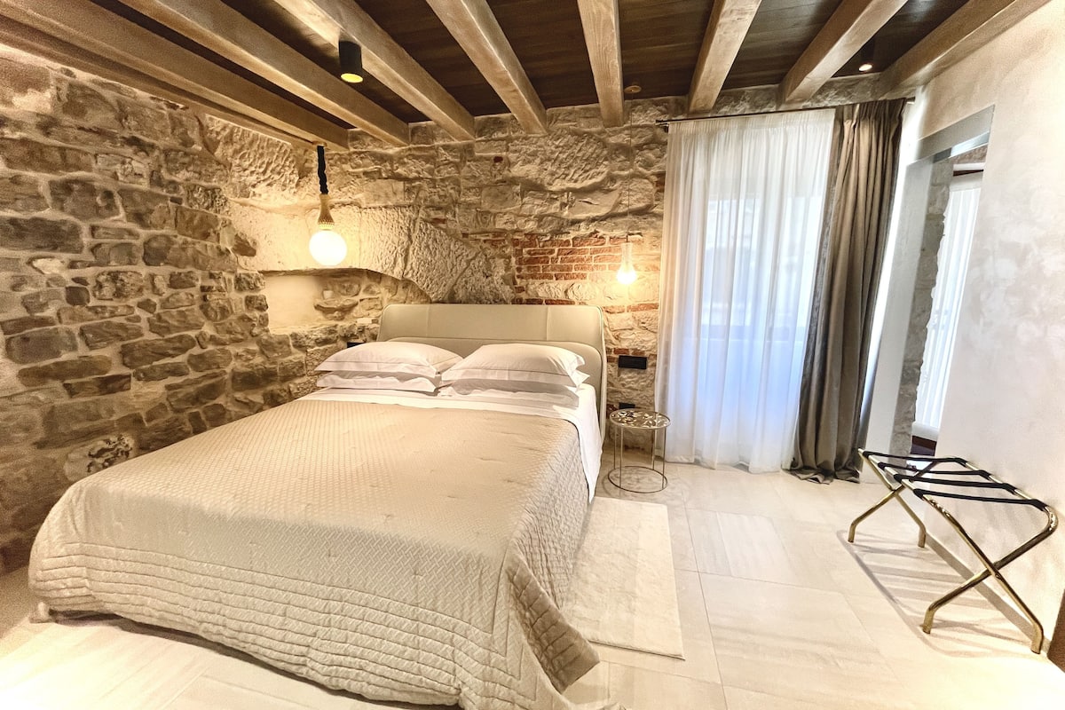 A bedroom with a stone wall, a bed with a white headboard, a bedside table, and a hanging light fixture.