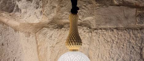 A hanging light fixture with a textured design, suspended from a wooden beam in a stone-walled room.