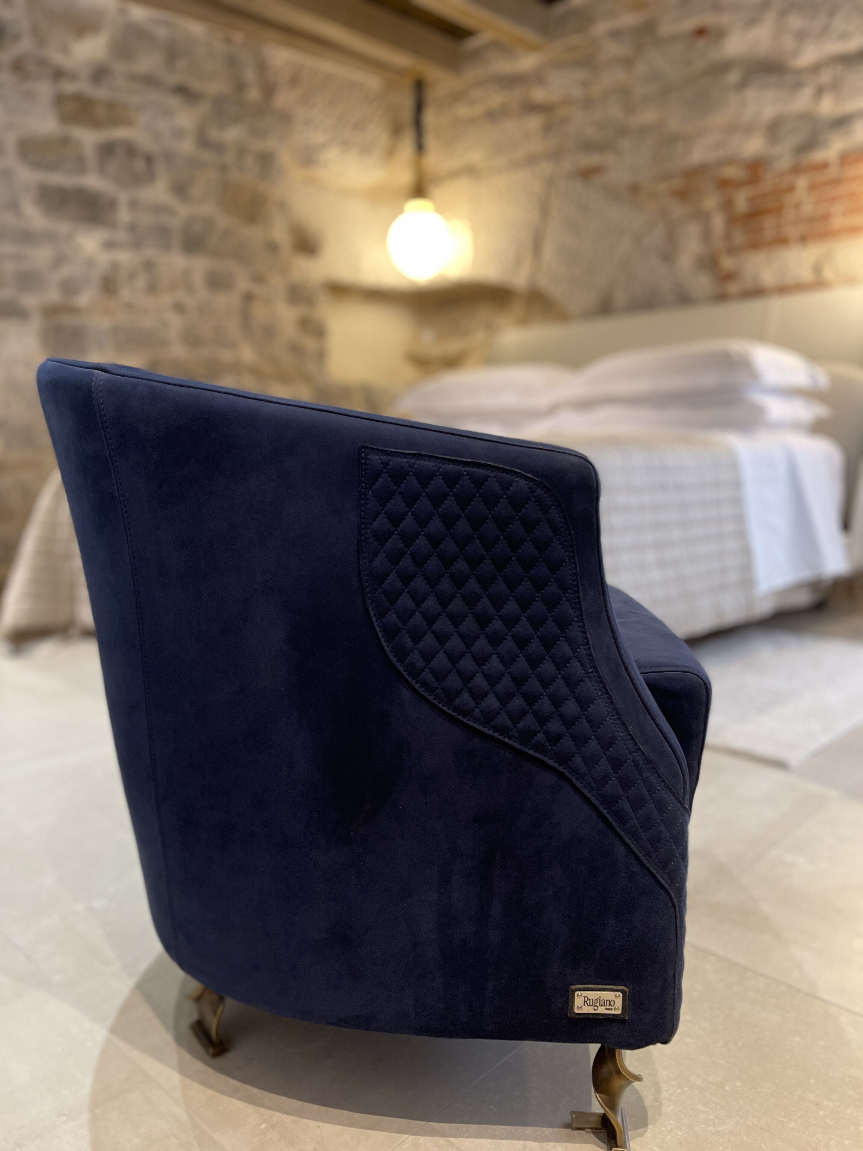 A dark blue upholstered chair with a quilted backrest and gold legs, placed on a tiled floor in a room with a stone wall and a bed in the background.