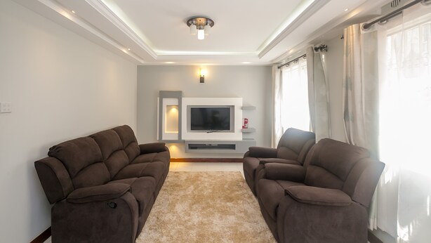 Un salon moderne avec deux canapés marron, un tapis beige, un téléviseur à écran plat et un luminaire suspendu au plafond.