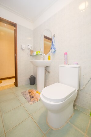A bathroom with a toilet, sink, and mirror. There is a trash bin and a mat on the floor.