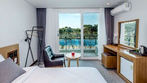 A hotel room with a bed, a desk, a chair, a small table with a red object, and a large window with a view of a pool and greenery.