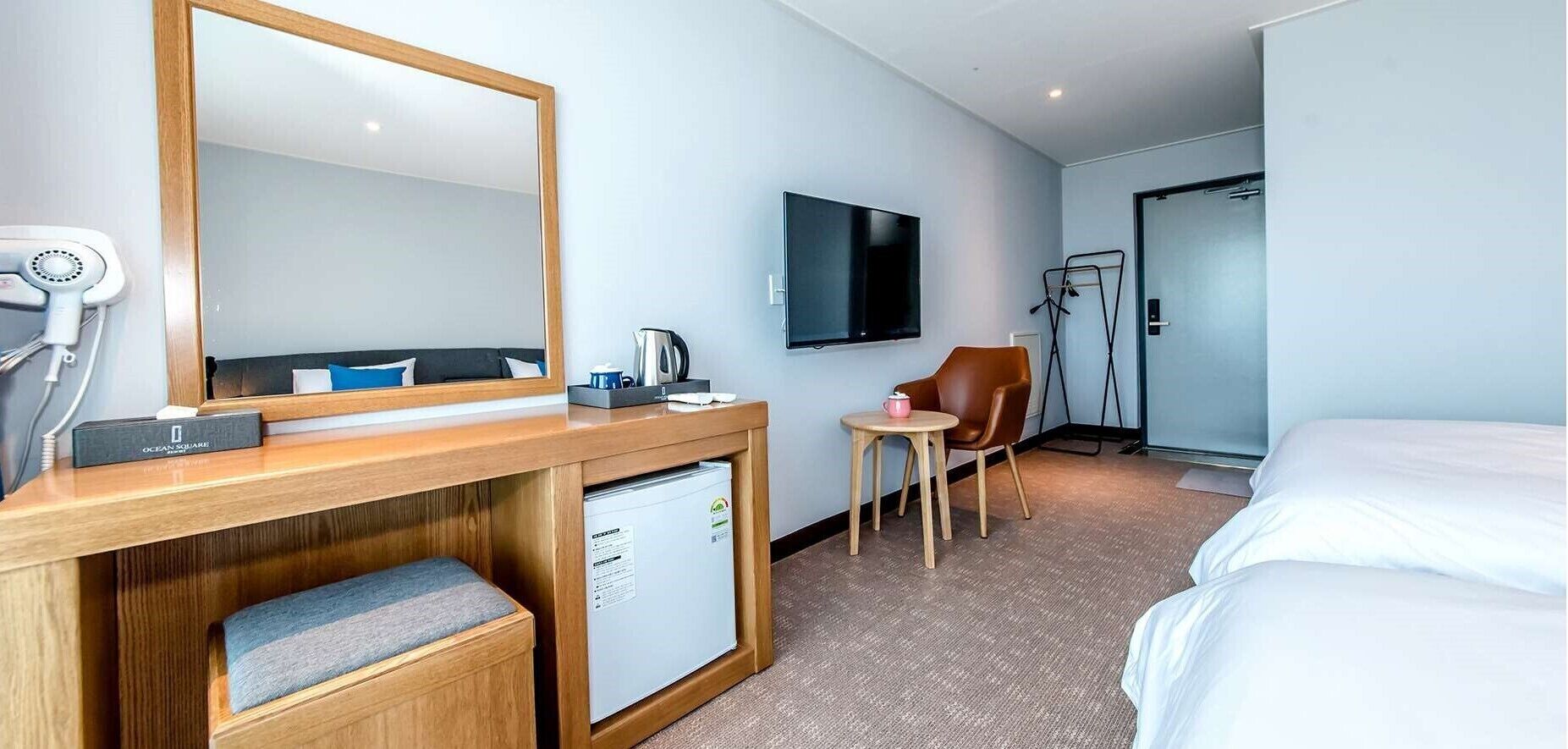 A hotel room with a bed, a desk with a mirror, a television, a chair, and a small table with a kettle and a cup.