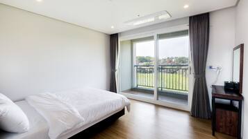 A bedroom with a bed, a wooden floor, a large window with curtains, and a balcony with a view of greenery.