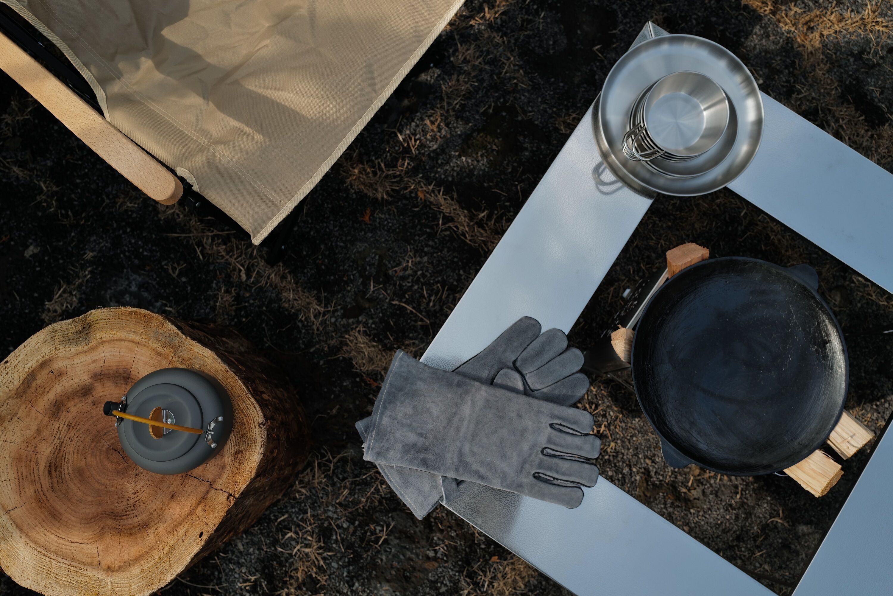 A camping setup with a table, a pot, a pair of gloves, a cup, and a lantern on a tree stump.