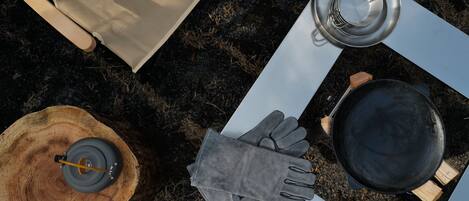 A camping setup with a table, a pot, a pair of gloves, a cup, and a lantern on a tree stump.