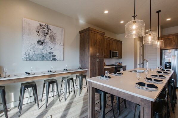 A modern kitchen with a long island, bar stools, wooden cabinets, stainless steel appliances, and pendant lighting.
