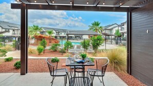 An outdoor patio with a table and chairs, a pergola with hanging lights, and a view of a residential area with houses and a swimming pool.