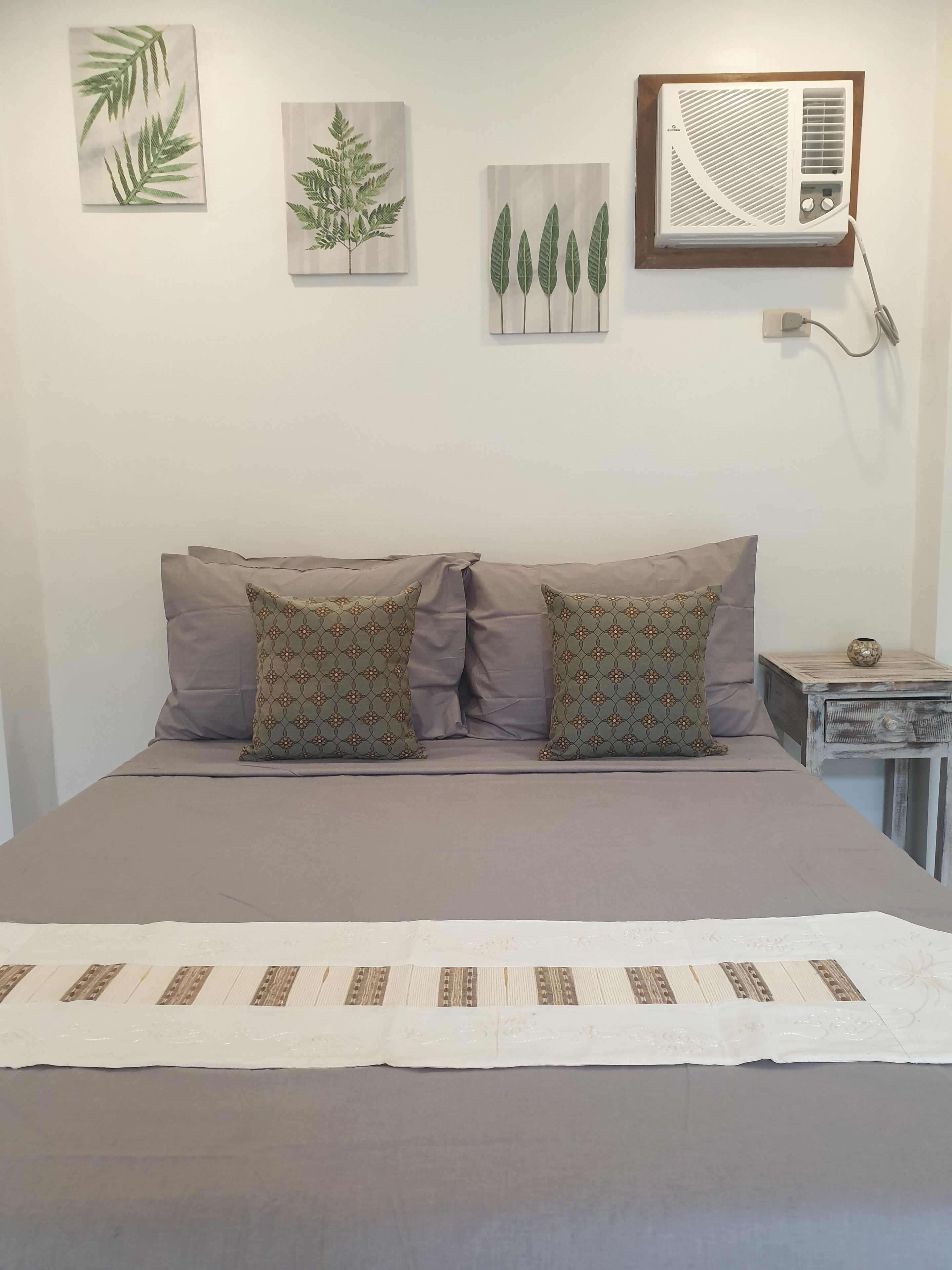 A bedroom with a bed, two patterned pillows, a wall-mounted air conditioner, and three leaf-patterned wall art pieces.