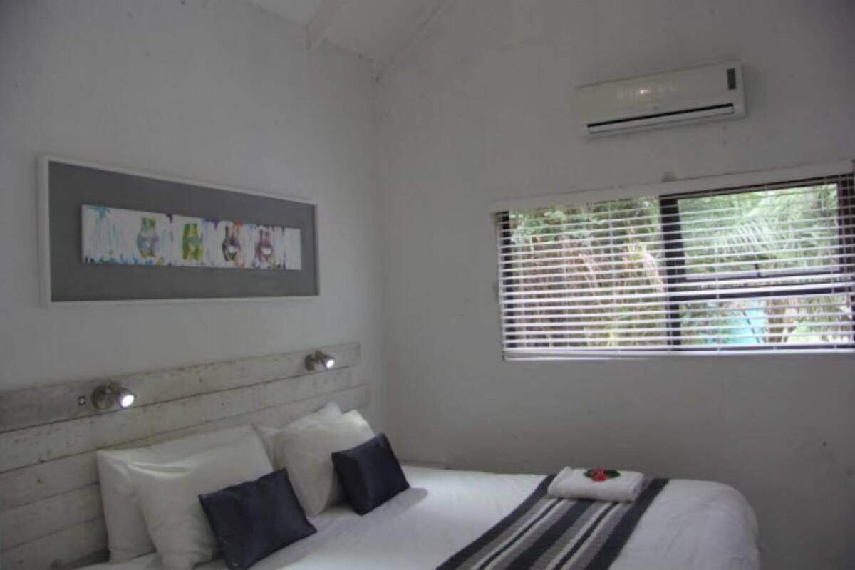 Une chambre à coucher avec un lit, des oreillers, une tête de lit, un climatiseur et une fenêtre avec des stores.