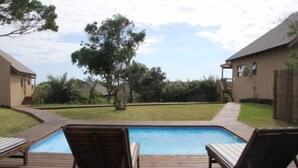 Uma piscina com deck de madeira e espreguiçadeiras, cercada por um quintal com árvores e uma casa ao fundo.