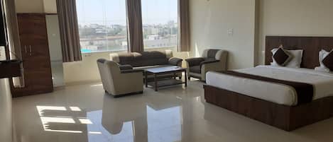 Una habitación de hotel con una cama, un sofá, una mesa de centro y una silla. Cuenta con un ventanal con cortinas, un ventilador de techo y un cuadro en la pared.