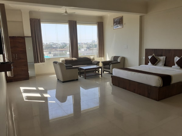 Una habitación de hotel con una cama, un sofá, una mesa de centro y una silla. Cuenta con un ventanal con cortinas, un ventilador de techo y un cuadro en la pared.