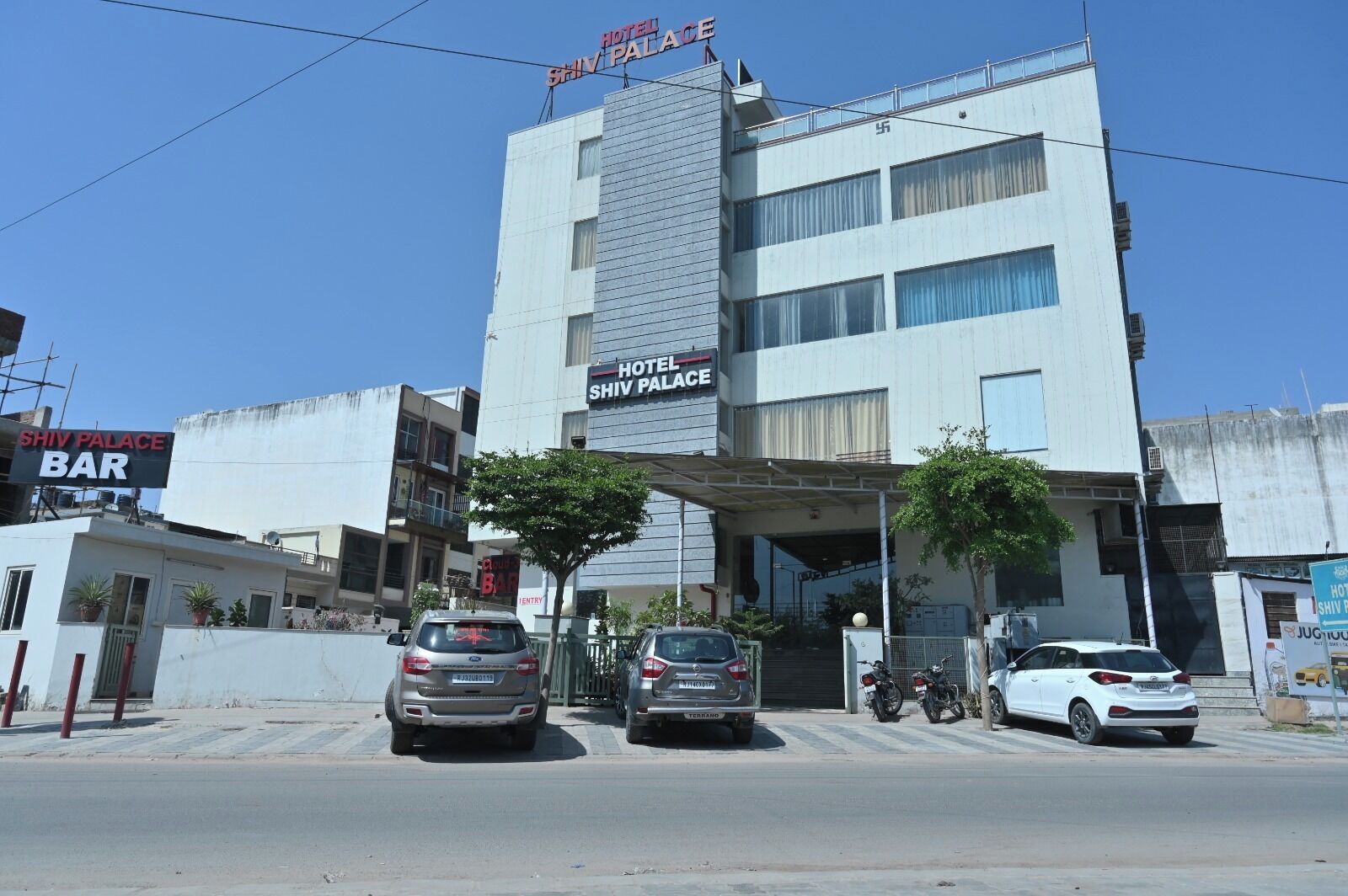 A hotel named Shiv Palace with a bar, multiple cars parked in front, and a clear blue sky.
