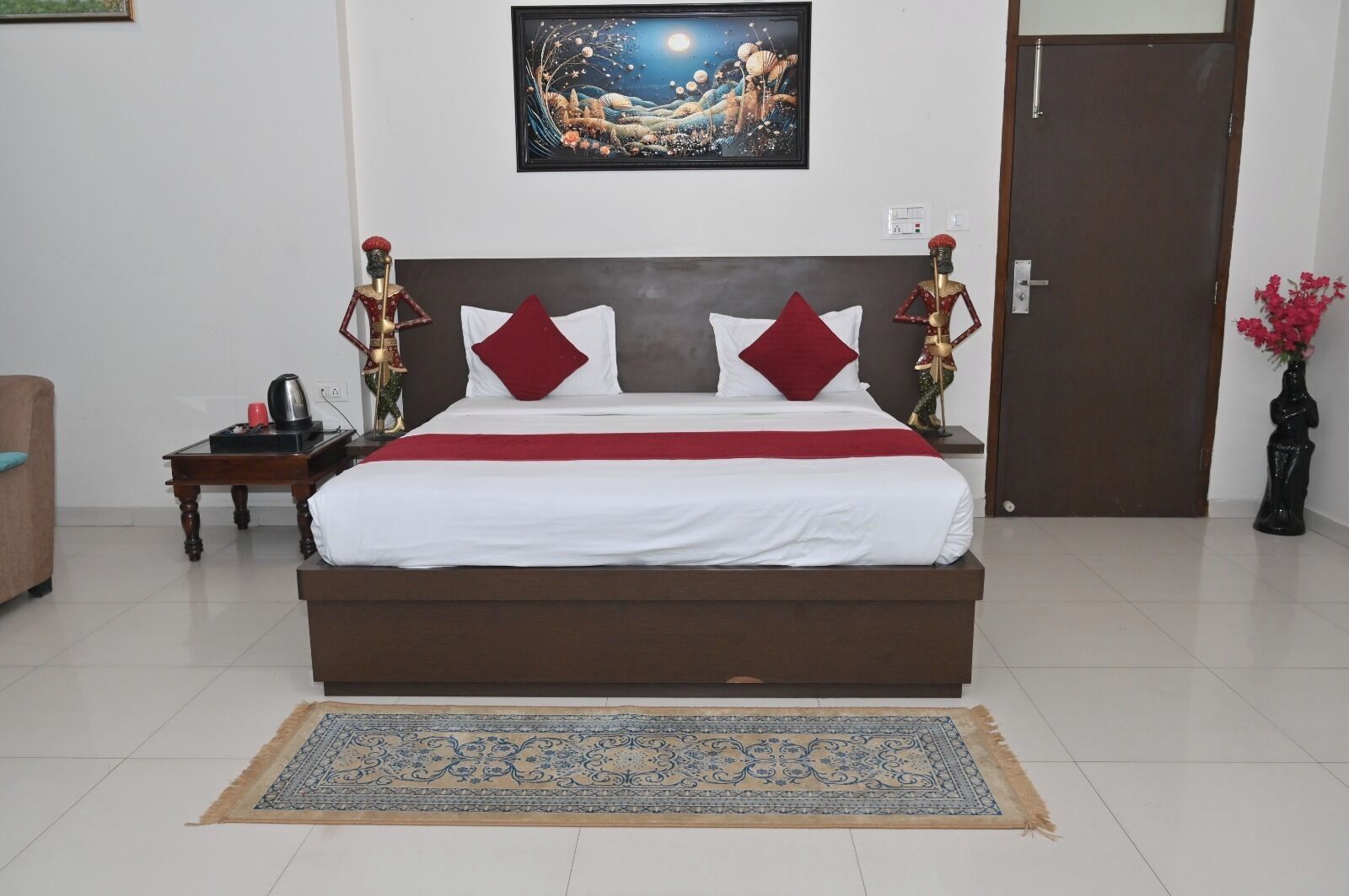 A neatly made bed with a red and white bedspread, two red pillows, and a decorative headboard. A nightstand with a kettle and a small plant is to the left. A framed artwork hangs on the wall above the bed.