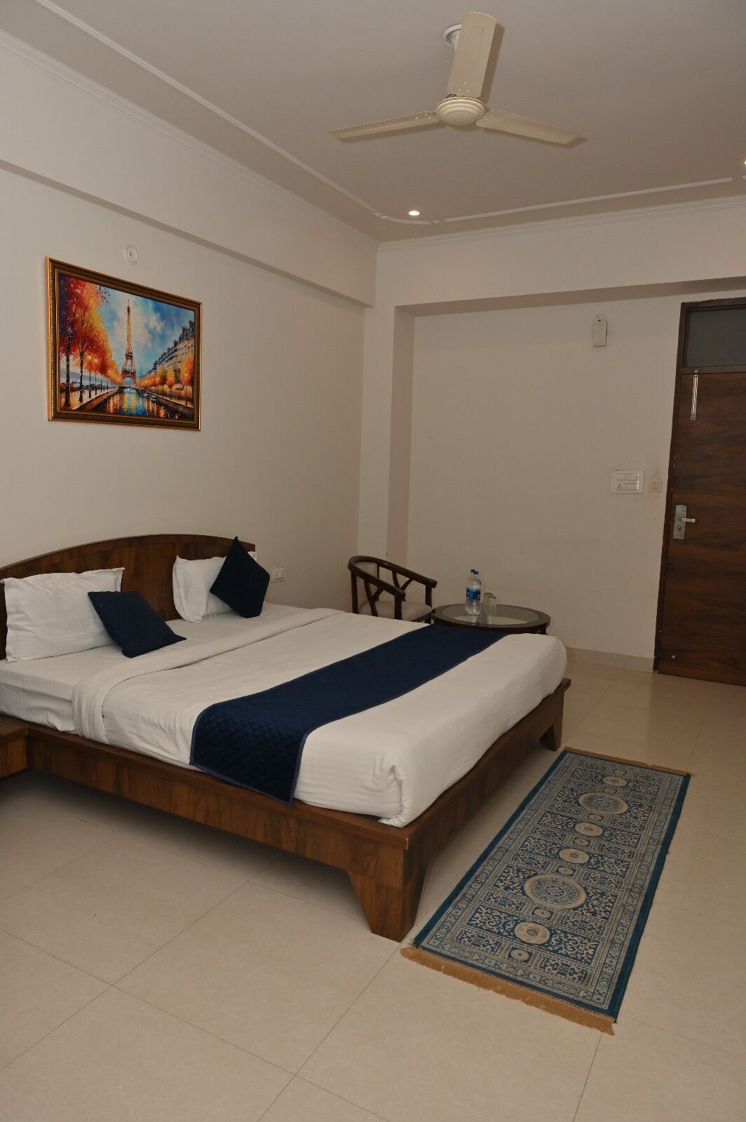 A hotel room with a bed, a ceiling fan, a small table with a bottle, a painting of a bridge, and a door.
