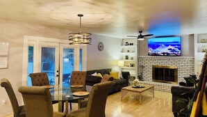 Een woonkamer met een open haard, een eetgedeelte met een glazen tafel, en een aan de muur bevestigde televisie.
