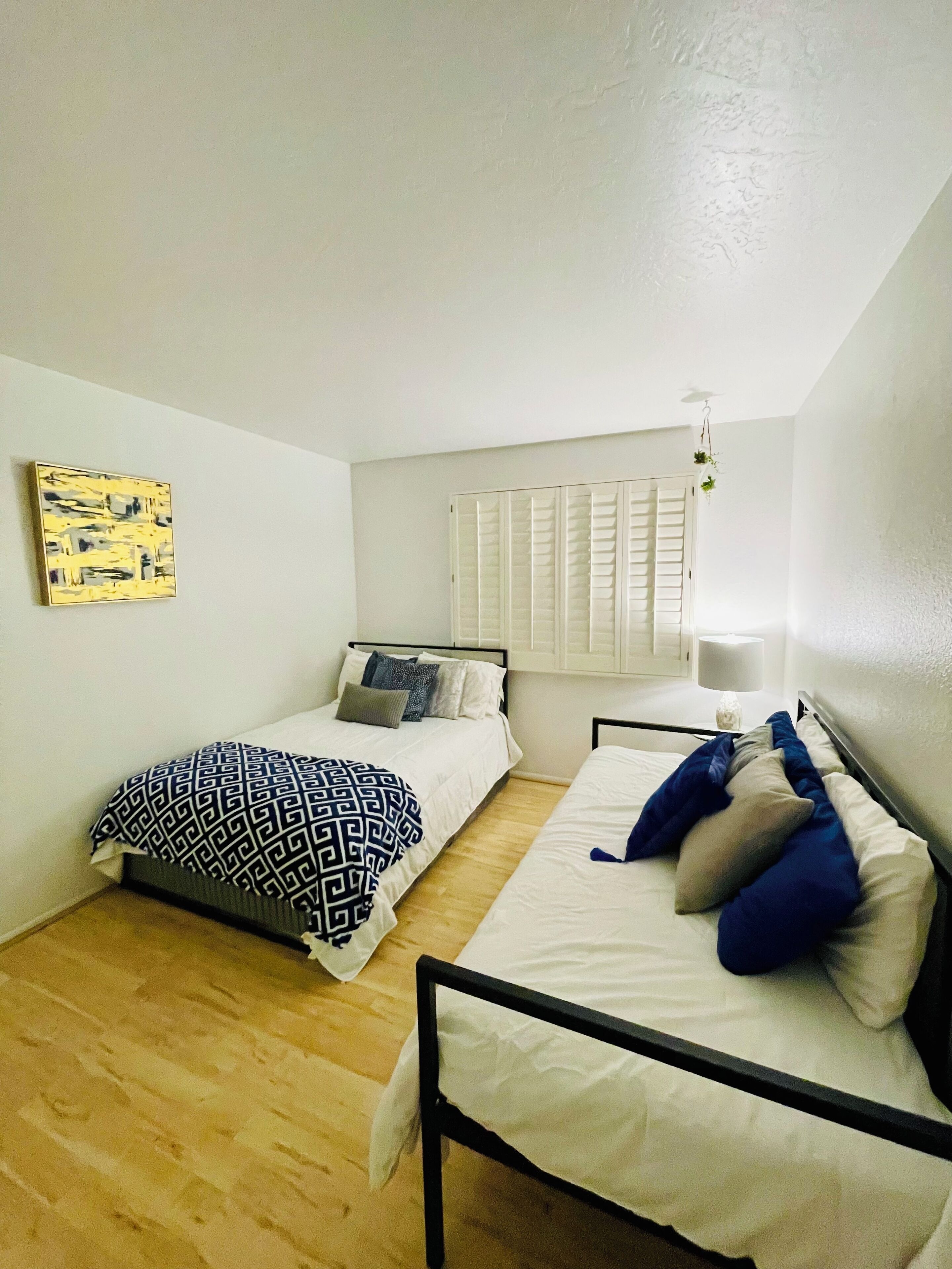 A room with two beds, a window with shutters, a hanging plant, and a painting on the wall.