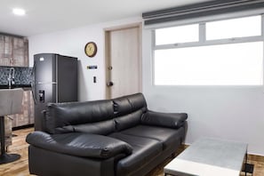 A modern living room with a black leather sofa, a small table, a refrigerator, a clock, and a window with blinds.