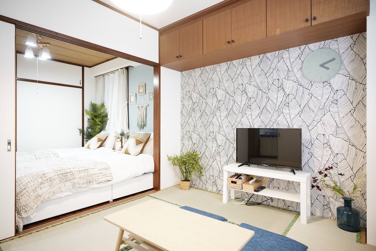 Un dormitorio con una cama, un televisor sobre un soporte blanco, un reloj en la pared y una planta.