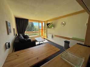 Una habitación con sofá, una mesa de madera, un televisor montado en la pared y un ventanal con vistas al exterior.