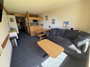 A compact living space with a sofa, a wooden table, a kitchenette, and a trash bin.