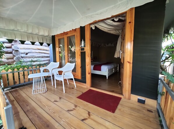 Une terrasse en bois avec un tapis rouge, des chaises en plastique blanches et un lit visible à travers une porte ouverte.