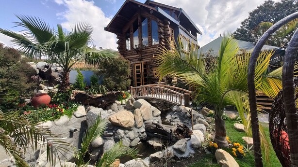 A rustic log cabin with a stone pathway leading to a wooden bridge, surrounded by lush greenery and a small waterfall. - Cabana Affair (Dehradun)