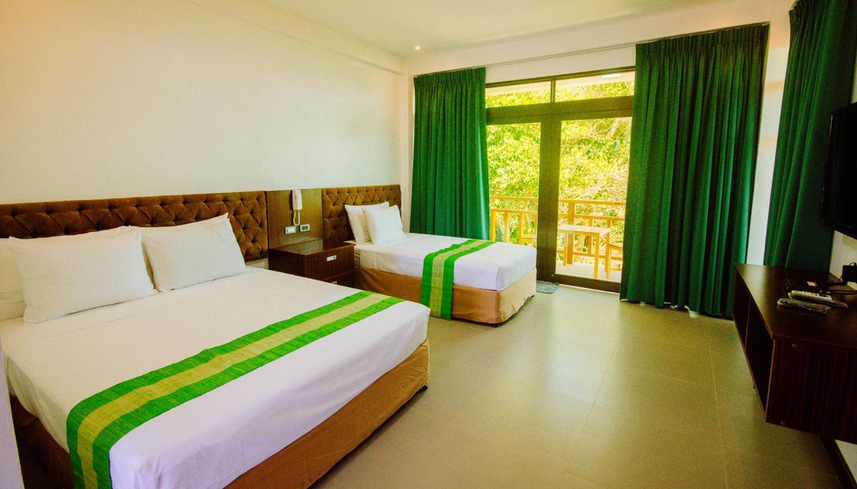 A hotel room with two beds, a wooden headboard, a desk with a television, and a balcony with green curtains.
