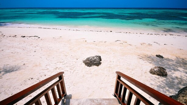 Una playa con agua turquesa cristalina y una orilla arenosa. Se observan barandillas de madera en primer plano.