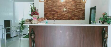 Un mostrador de recepción con un panel de madera, una barandilla metálica y elementos decorativos como un jarrón con flores y una estatua.