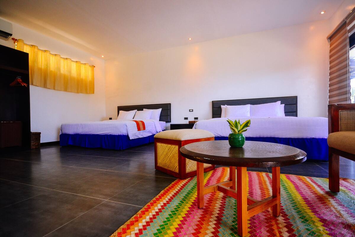 A hotel room with two beds, a small round table, a colorful rug, and a window with blinds.