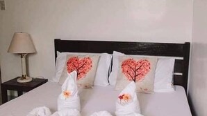 Una cama bien hecha con dos toallas dobladas en forma de corazón, una lámpara en una mesita de noche y dos almohadas con diseño de corazón.