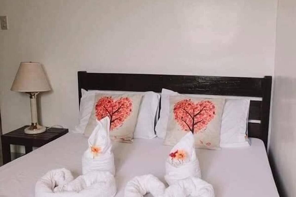 Ein ordentlich gemachtes Bett mit zwei Handtüchern, die zu Herzchen geformt sind, einer Lampe auf einem Nachttisch und zwei Kissen mit Herzmuster.