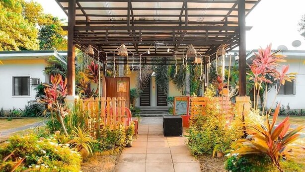 Un sendero cubierto en un jardín que lleva a la entrada de un edificio, rodeado de plantas tropicales y una valla de madera.