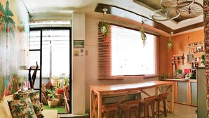 Un salon chaleureux avec une table et des chaises en bois, un canapé agrémenté de coussins aux motifs tropicaux et un coin cuisine équipé d’un réfrigérateur et d’étagères.