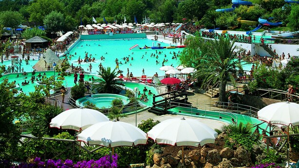 A large outdoor swimming pool surrounded by beach chairs and umbrellas, with a view of mountains in the background. - Bungalow Village Le Caravelle - by Mamberto S.r.l. (Ceriale)