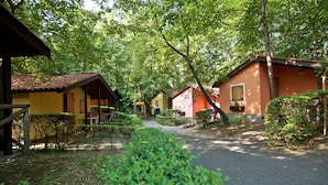 A row of small, single-story cabins with a paved path leading through them, surrounded by lush greenery. - Bungalow Village Le Caravelle - by Mamberto S.r.l. (Ceriale)
