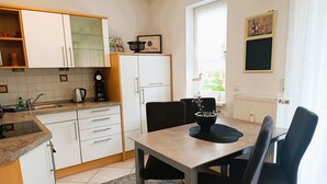 Et moderne køkken med hvide skabe, et spisebord med stole, en kaffemaskine og et vindue med udsigt til grøn bevoksning.